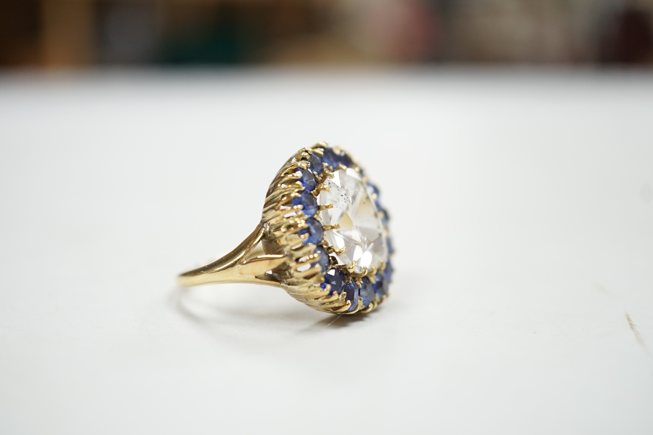 An early 20th century yellow metal and single stone cushion cut white sapphire set dress ring, bordered by round and oval cut blue sapphires, size P, gross weight 9.4 grams.